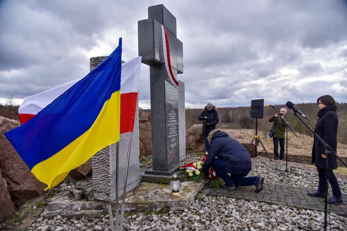 Ludzie składają kwiaty pod kamiennym pomnikiem w kształcie krzyża, przy którym stoją polska i ukraińska flaga; w tle widać mikrofony i osoby uczestniczące w uroczystości upamiętniającej.