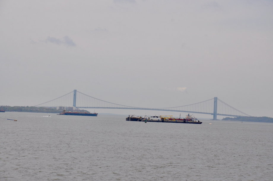 W oddali widoczny dwupoziomowy most Ver­ra­za­no–Nar­rows, łączący Brooklyn ze Staten Island. Najdłuższe przęsło ma długość blisko 1300 m. Za mostem rozpościera się Ocean Atlantycki. Giovanni da Verrazzano w 1524 roku stał na czele wyprawy, która odkryła tereny dzisiejszego Nowego Jorku, wówczas zamieszkiwane przez Indian. 