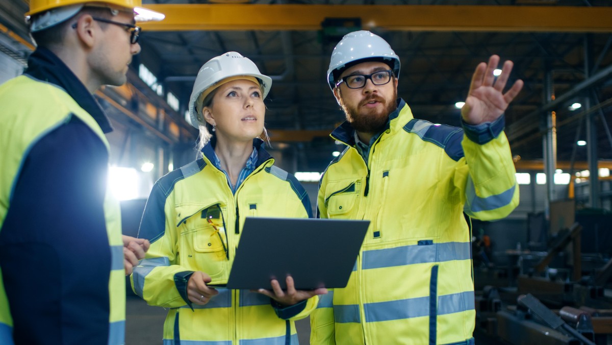 Male and Female Industrial Engineers Talk with Factory Worker while Using Laptop. They Work at the Heavy Industry Manufacturing Facility.