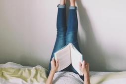 Woman Lying On Bed Reading Book With Legs Raised