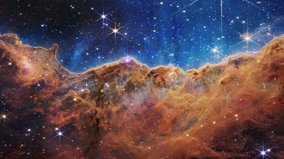 Ten krajobraz "gór" i "dolin" usianych gwiazdami to w rzeczywistości krawędź pobliskiego, młodego regionu gwiazdotwórczego o nazwie NGC 3324 w Mgławicy Carina. Uchwycony w świetle podczerwonym przez Kosmiczny Teleskop Jamesa Webba, obraz ten po raz pierwszy ujawnia niewidoczne wcześniej obszary narodzin gwiazd. Źródło: NASA, ESA, CSA, STScI
