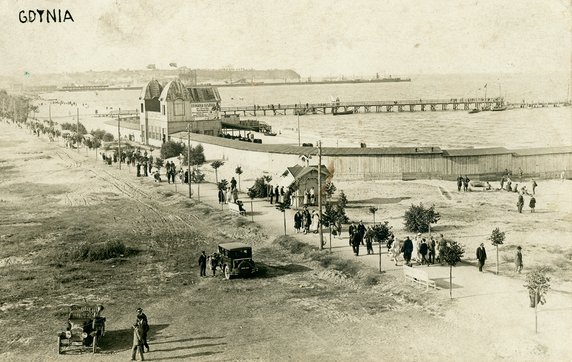 Zakłady Kąpielowe w Gdyni w 1925 r. Na plaży przy promenadzie widoczny ciąg drewnianych zabudowań przebieralni przystających do głównego, wysokiego budynku. Na jego ścianie bocznej mieści się reklama „DROGERJI KASZUBJA”. Nieco dalej widoczny drewniany pomost pasażerski, a w tle znajduje się port tymczasowy oraz Kępa Oksywska