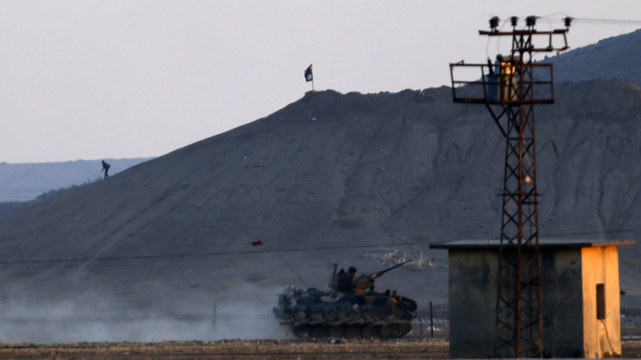 An ISIS fighter walks near an ISIS flag as a Turkish army vehicle takes position near the Syrian town of Kobani.