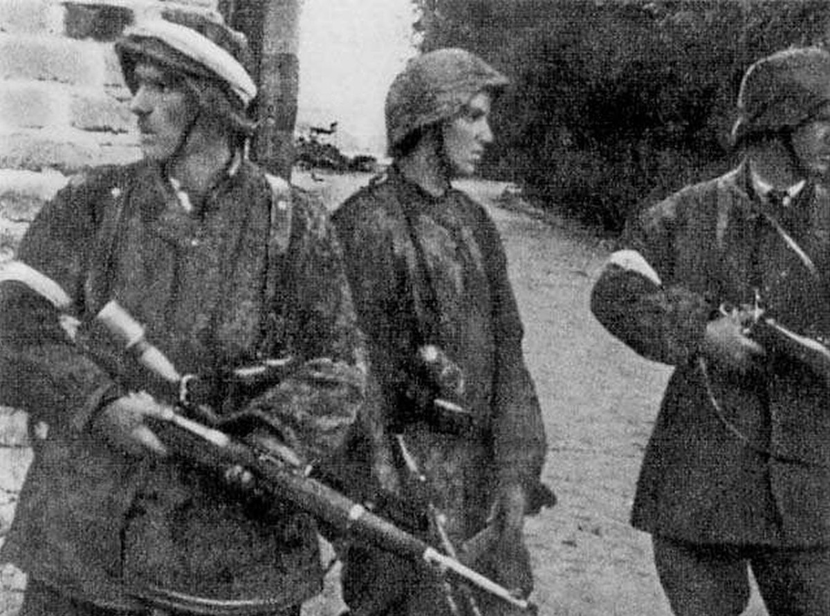 Patrol z II plutonu "Alek" 2. kompanii "Rudy" batalionu "Zośka" 5 sierpnia 1944 r. na Gęsiówce. Od lewej stoją: Wojciech Omyła "Wojtek", Juliusz Bogdan Deczkowski "Laudański" i Tadeusz Milewski "Ćwik". Milewski poległ na terenie "Gęsiówki" w dniu zrobienia tego zdjęcia; Omyła zginie trzy dni później. W chwilę po zrobieniu tego zdjęcia, w mur nad głowami powstańców uderzył niemiecki pocisk karabinowy - nikomu nic się nie stało