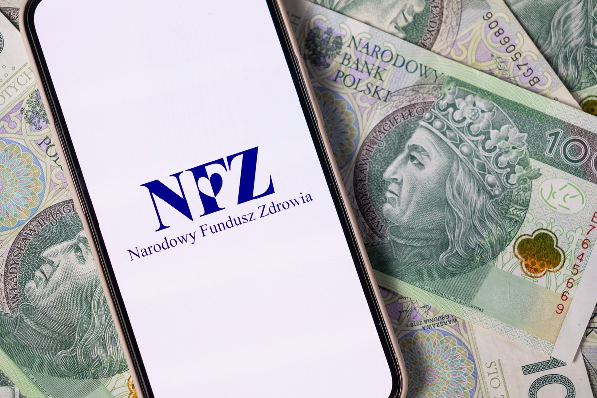 Smartfon z napisem NFZ na tle polskich banknotów 100 zł. NFZ odbiera prawo do leczenia po 30 dniach od ustania ubezpieczenia