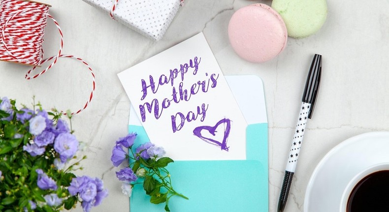 Happy Mother's Day card beside pen, macaroons, flowers, and gift box near a coffee cup uon a saucer [Photo: George Dolgikh]