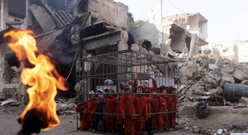 An activist (not pictured) holds a burning torch near children carrying banners inside a cage during a protest against forces loyal to Syria's President Bashar al-Assad in Douma, eastern Al-Ghouta, near Damascus, February 15, 2015. The protest, which made children wear orange suits depicting victims of the Islamic State, calls to compare forces loyal to Syria's president Bashar al-Assad to Islamic State forces, and to draw attention to residents living under siege and dying from strikes by forces loyal to Syria's president Bashar Al-Assad, activists said.