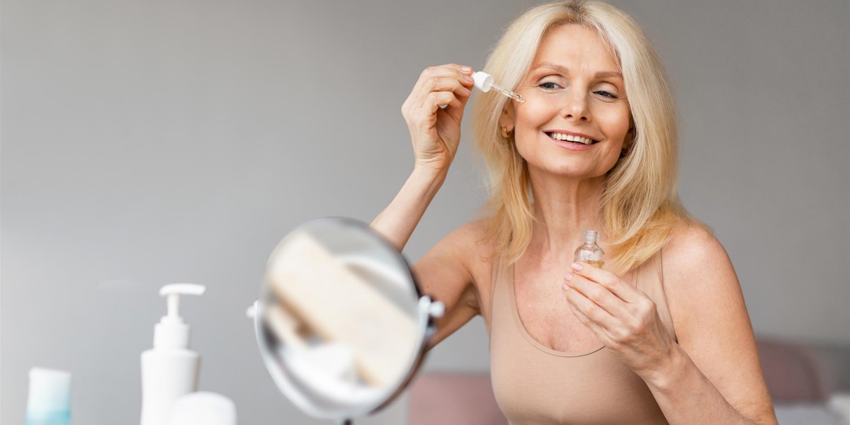 Beautiful mature woman applying eye serum with dropper and smiling at mirror, holding bottle with be