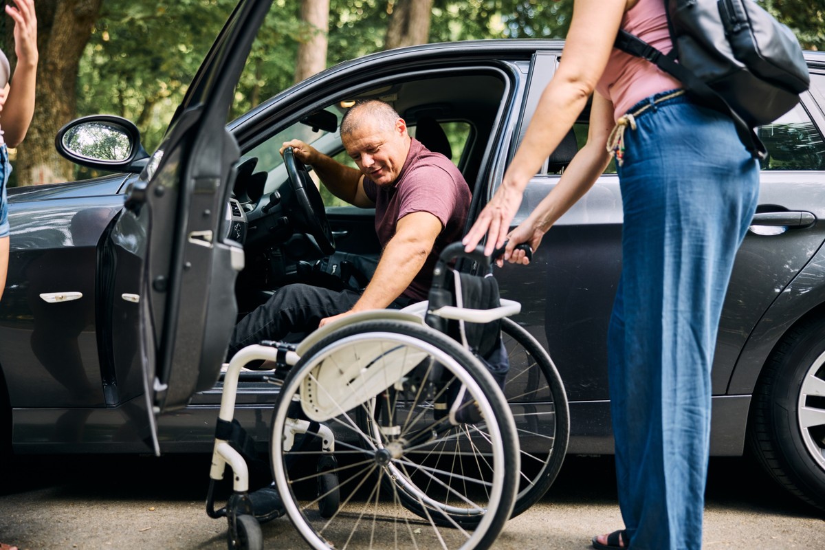 Osoba z niepełnosprawnością wychodzi z samochodu, wspiera ją osoba trzymająca wózek inwalidzki
