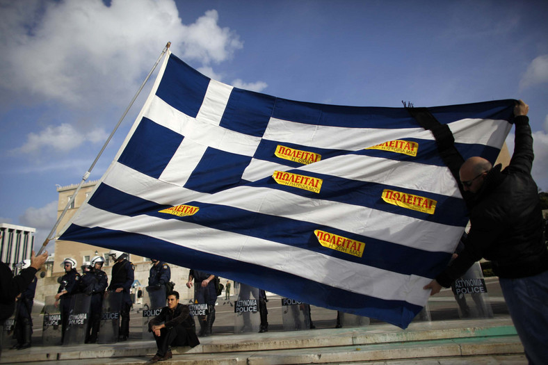 Protestujący Grecy z flagą swojego kraju obklejoną kartkami z napisem 