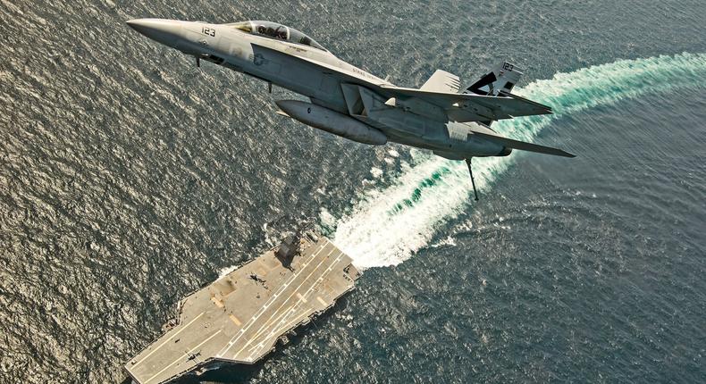 An F/A-18F Super Hornet jet flies over the USS Gerald R. Ford as the U.S. Navy aircraft carrier tests its new launch and flight arrest systems