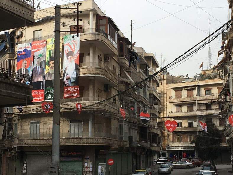 Widok na budynku w centrum miasta - widoczne plakaty z podobiznami przywódców Rosji, Iranu, Hezbollahu i dekoracje z barwami Syrii i wizerunkiem prezydenta al-Asada