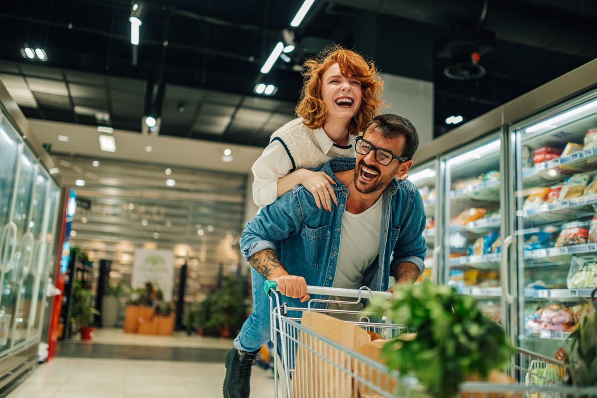 Mężczyzna pcha wózek sklepowy w supermarkecie, na jego plecach siedzi uśmiechnięta kobieta. Para dobrze się bawi się podczas zakupów w dziale z chłodziarkami.
