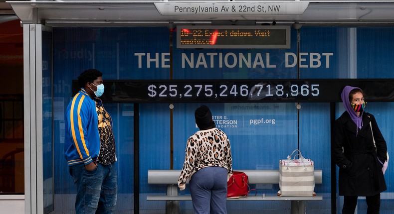 Passengers wearing face masks wait for their bus in front of a national debt display on Pennsylvania Ave. NW in Washington on Monday, May 18, 2020
