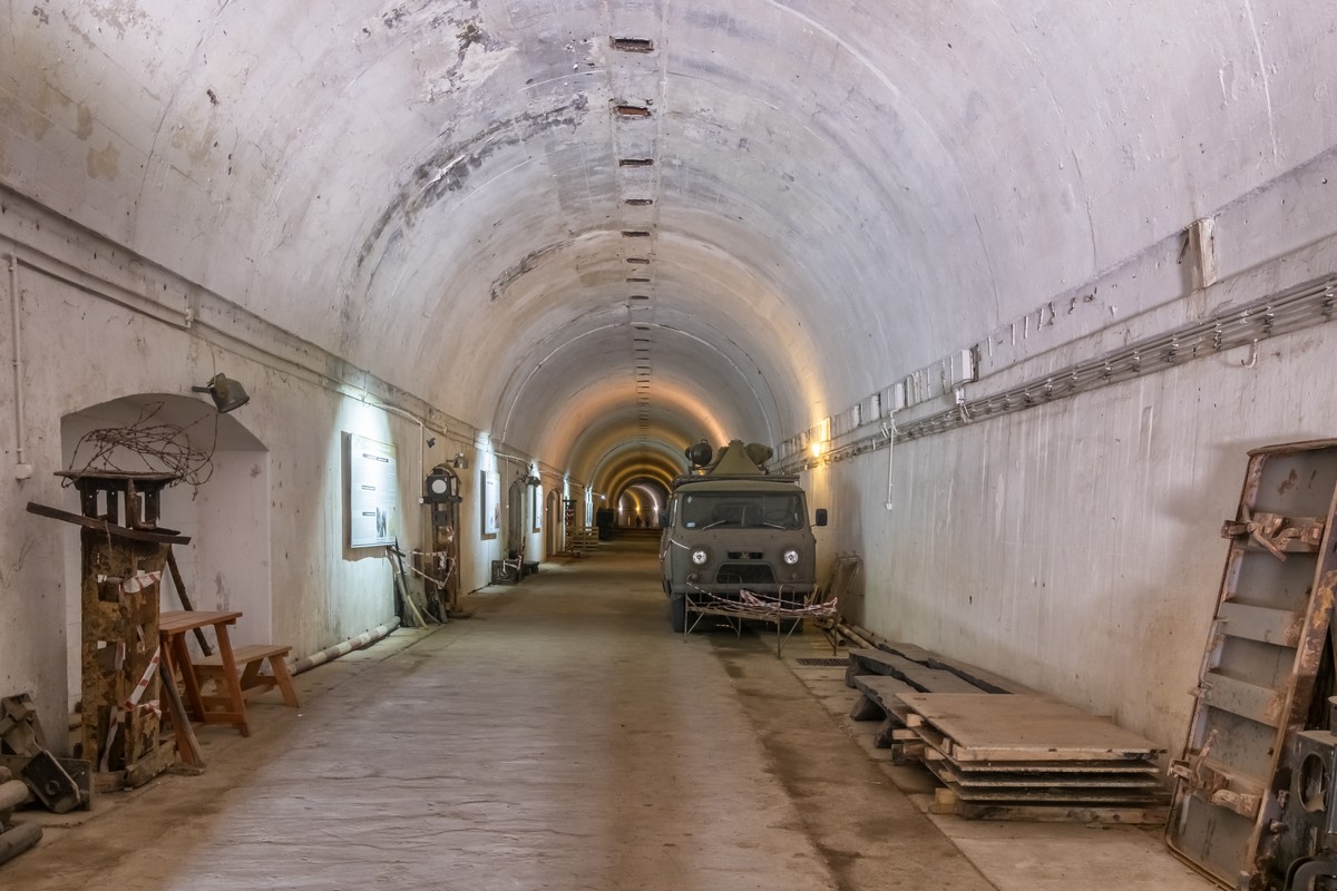 Wnętrze betonowego tunelu schronu przeciwlotniczego z widocznym zaparkowanym wojskowym pojazdem, oświetlone bocznymi lampami, z elementami wyposażenia i materiałami budowlanymi ustawionymi przy ścianach