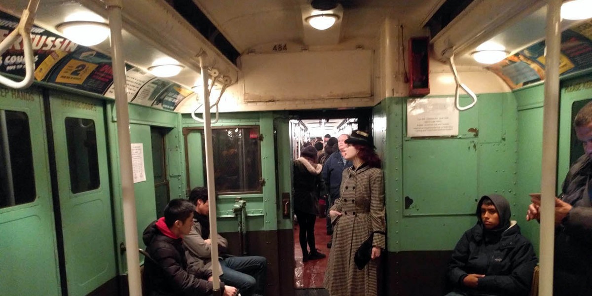 A woman dressed in early 20th-century fashion (while listening to music on her 21st-century smartphone) stands on the annual Shopper's Special subway line.