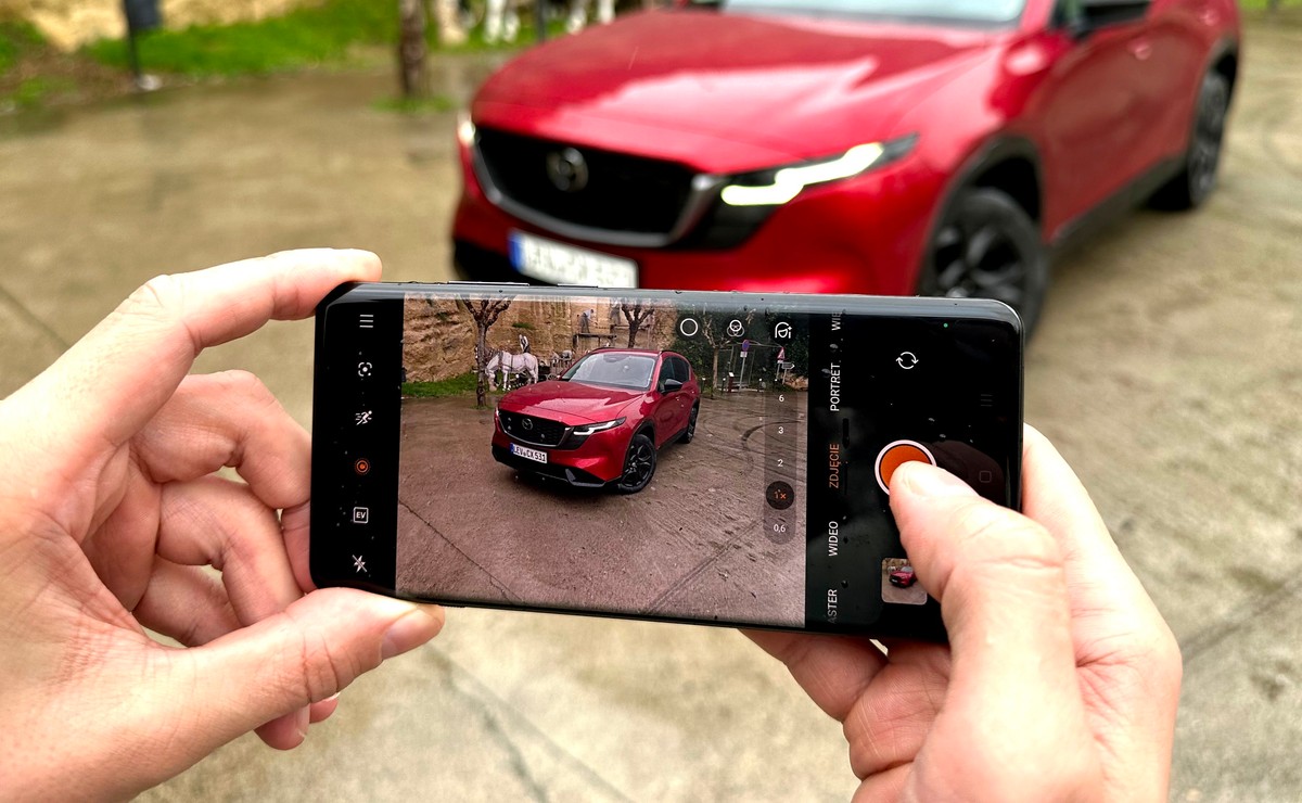 Osoba trzymająca smartfon i robiąca zdjęcie czerwonego samochodu na parkingu, na ekranie telefonu widoczne menu aparatu w języku polskim, ilustracja do artykułu o kontrolach abonamentu RTV w autach