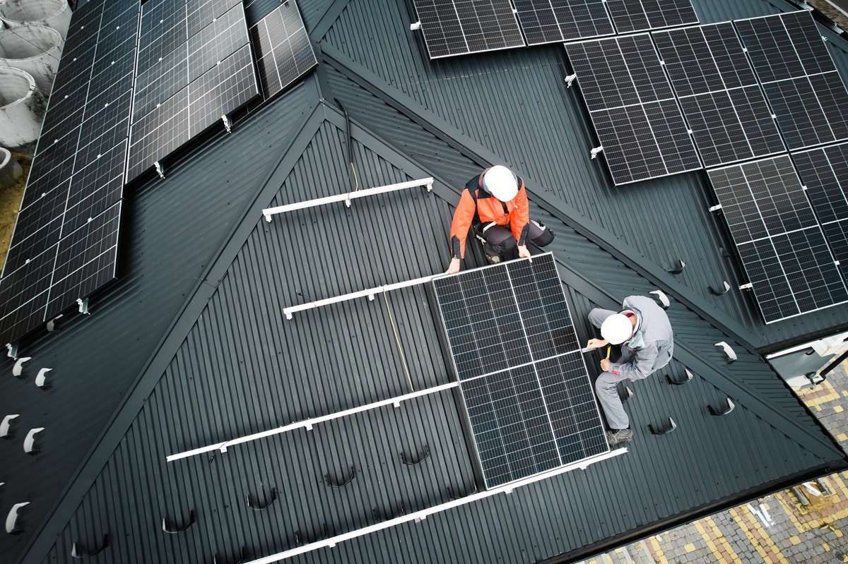 Dwóch monterów w kaskach ochronnych instaluje panel fotowoltaiczny na spadzistym dachu budynku. Wokół widoczne są już zamontowane moduły solarne, a pracownicy mocują kolejny element konstrukcji.