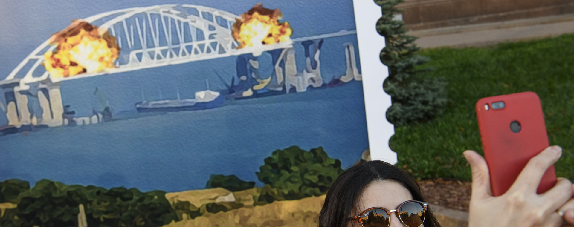 Ludzie pozują do zdjęć i robią selfie przed dużym plakatem znaczka pocztowego, przedstawiającym płonący Most Krymski. Kijów, Ukraina. 8 października 2022 r.