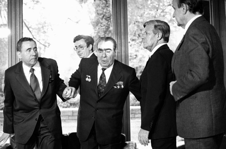 Od lewej: minister spraw zagranicznych ZSRR Andriej Gromyko i kanclerz Niemiec Zachodnich Helmut Schmidt pomagają Breżniewowi stanąć na nogi, gdy ten nie był w stanie podnieść się z kanapy bez pomocy.