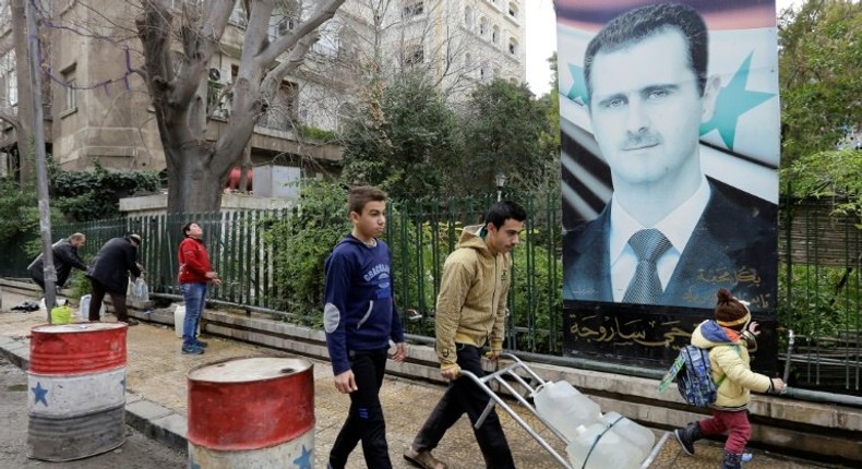 Syrians walk past a poster of President Bashar al-Assad as they go to fill plastic containers with water at a public fountain in the capital Damascus