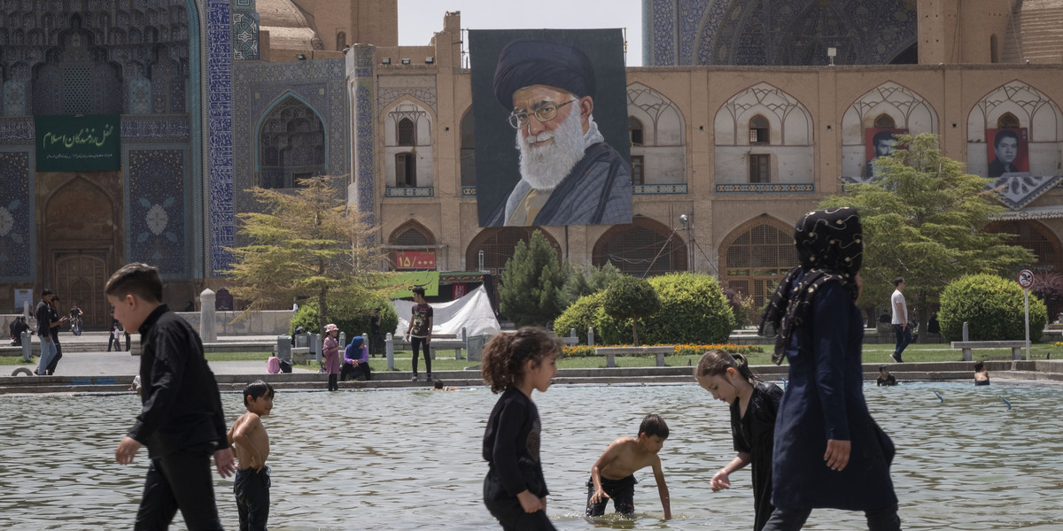 Irańskie dzieci bawiące się w miejskiej fontannie. W tle portret, na którym widoczny jest przywódca Iranu Ali Chamenei.
