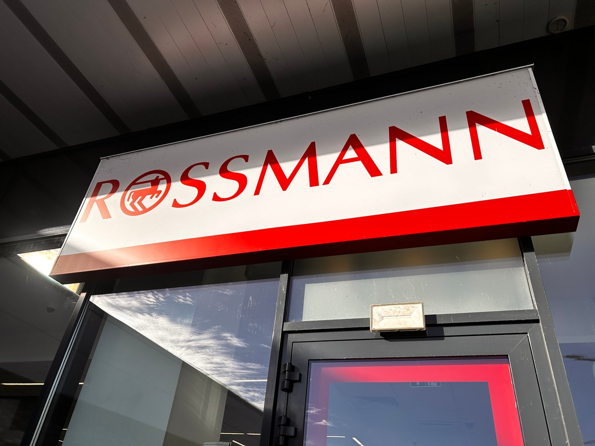 Szyld sklepu Rossmann z czerwonym logo i nazwą sieci na białym tle, umieszczony nad wejściem do drogerii.