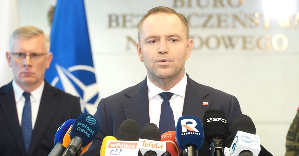 Prezydent Karol Nawrocki podczas konferencji prasowej w Biurze Bezpieczeństwa Narodowego, stojący za mównicą z mikrofonami mediów, obok szefa BBN Sławomira Cenckiewicza, w tle widoczna flaga NATO i napis „Biuro Bezpieczeństwa Narodowego”