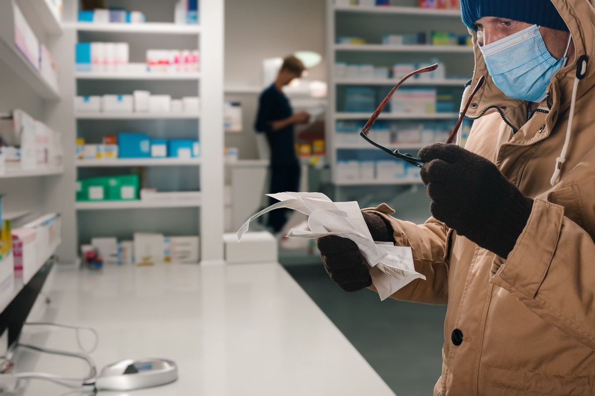 Osoba w maseczce ochronnej i rękawiczkach trzyma recepty i okulary w aptece, w tle farmaceuta przy półkach z lekami.