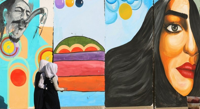 Iraqi women walk past a mural inspired by ongoing anti-government protests in Tahrir square in Baghdad while political leaders are struggling to choose a new prime minister