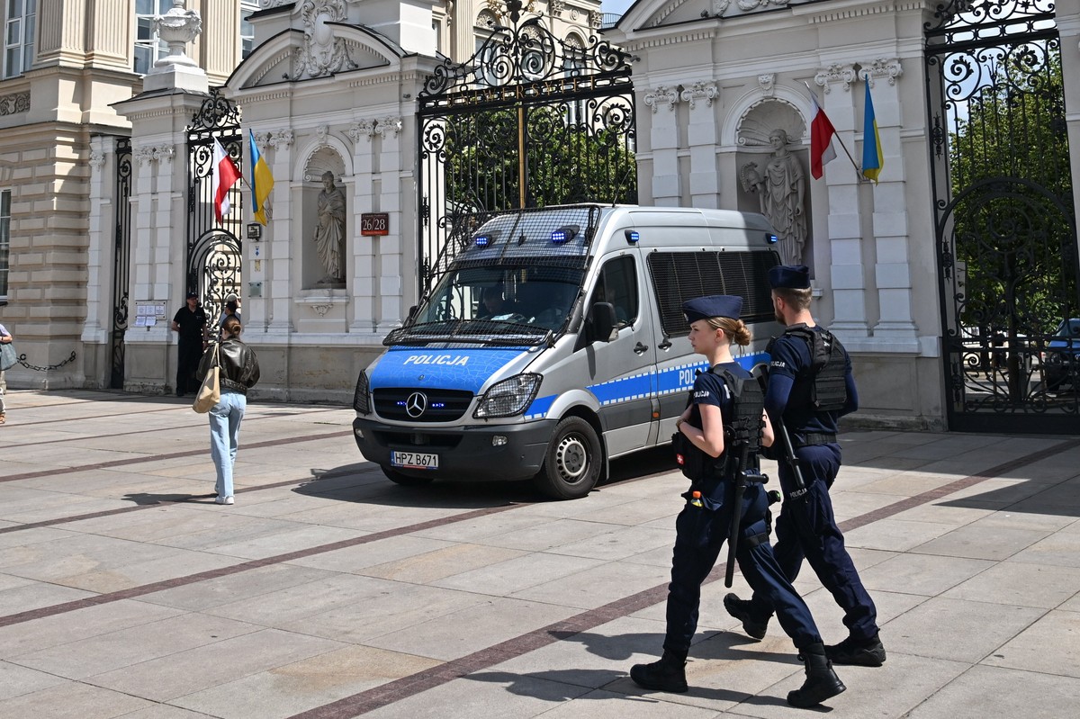 Policja w mundurach idzie chodnikiem obok radiowozu zaparkowanego przy bramie Uniwersytetu Warszawskiego; w tle przechodnie i flagi państwowe. Mundurowi otrzymają nowy dodatek mieszkaniowy