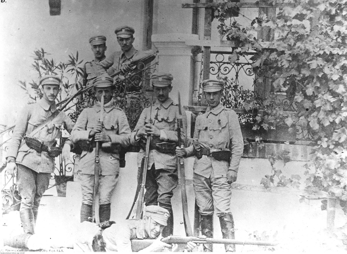 Patrol strzelecki. Na schodach stoją: Ludwik Kmicic-Skrzyński (z lewej), Władysław Belina-Prażmowski (z prawej). Niżej od lewej stoją: Zygmunt Bończa-Karwacki, Stanisław Grzmot-Skotnicki, Janusz Głuchowski, Antoni Jabłoński; leży Stefan Hanka-Kulesza celujący z karabinu Mannlicher wz. 1890. Data wydarzenia: 1914