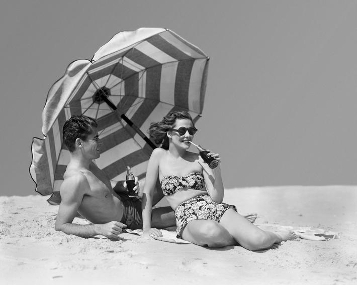1950s COUPLE MAN WOMAN SUNBATHING DRINKING BOTTLES SODA BY BEACH UMBRELLA