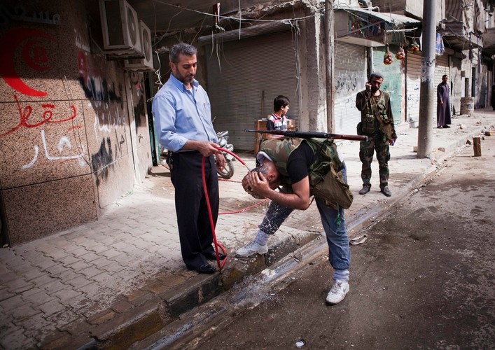 Bojownik myje się pod strumieniem wody z węża trzymanego przez jednego z mieszkańców zrujnowanego miasta