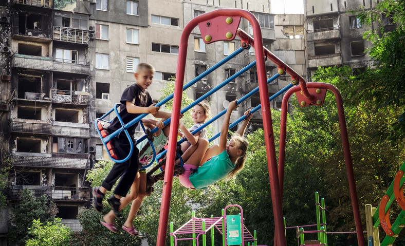 Dzieci na placu zabaw, w tle ostrzelany budynek w Mariupolu