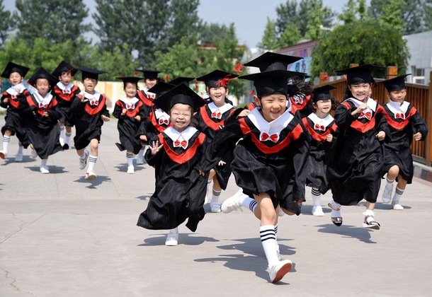 Children in gowns and mortarboards run with smiles during their kindergarten graduation ceremony in 