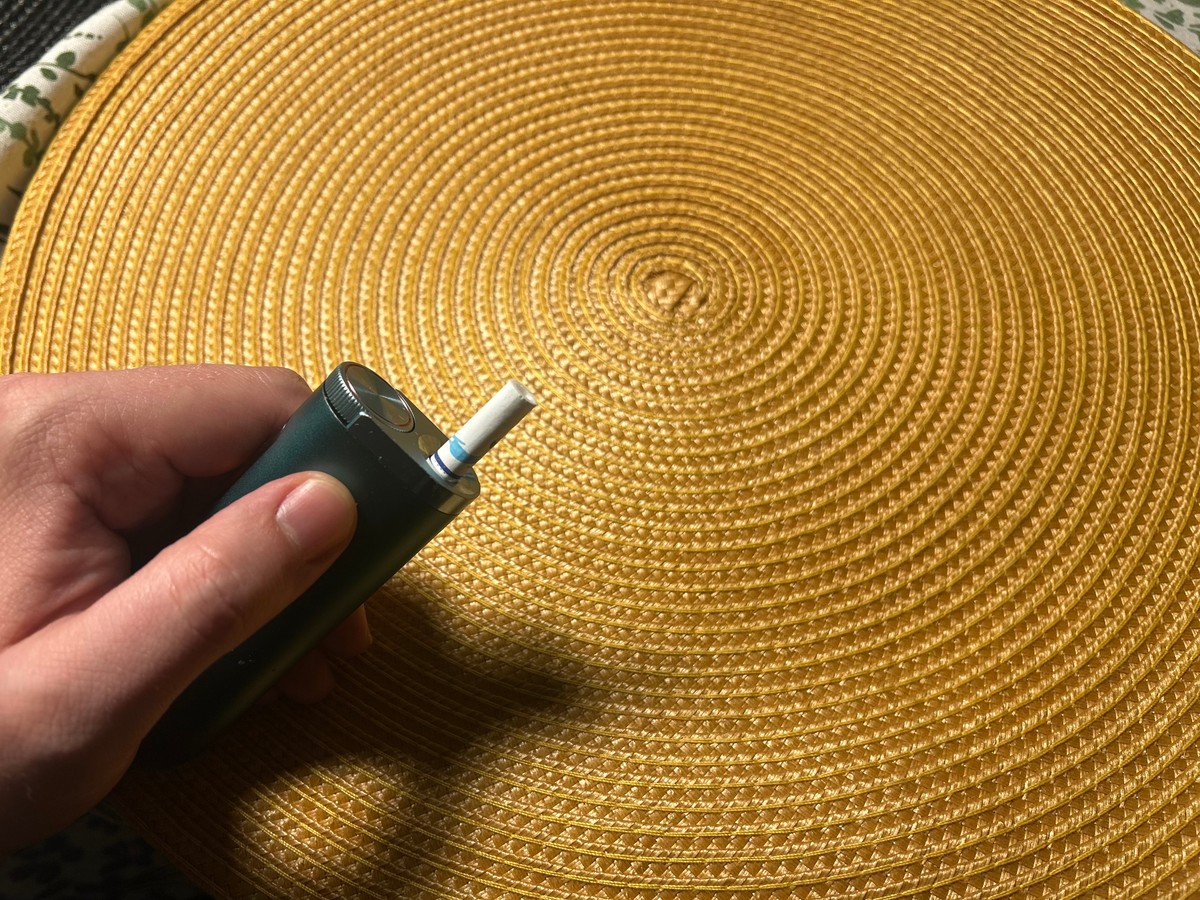 Dłoń trzymająca podgrzewacz tytoniu z włożonym wkładem tytoniowym, widziana z bliska na tle okrągłej, plecionej, złotej podkładki leżącej na stole.