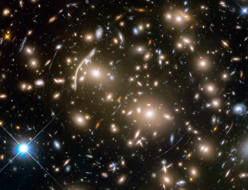 Gromada galaktyk Abell 370. Należące do niej galaktyki są ze sobą związane grawitacyjnie. Wyraźnie widać tajemnicze, niebieskie łuki światła. Astronomowie nie od razu wiedzieli, czym one są. Okazało się, że to tak zwane artefakty grawitacyjne, czyli silnie zniekształcone obrazy odległych galaktyk, położonych – patrząc z naszego punktu widzenia – daleko za samą gromadą. Te odległe galaktyki są tak słabo widoczne, że bezpośrednio nie jest w stanie zaobserwować ich nawet Teleskop Hubble'a. Pozwala na to dopiero "kosmiczna lupa”, czyli ogromna masa pomiędzy nimi a obserwatorem na Ziemi.