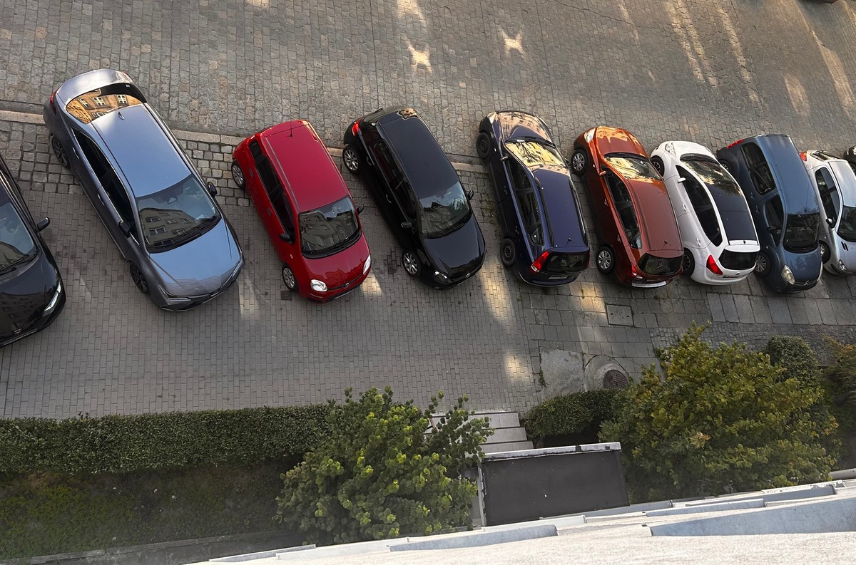 Rząd samochodów zaparkowanych wzdłuż chodnika przy bloku mieszkalnym, widok z góry; zdjęcie ilustruje temat minimalnej liczby miejsc postojowych w inwestycjach mieszkaniowych po zmianach przepisów od sierpnia 2025 roku.