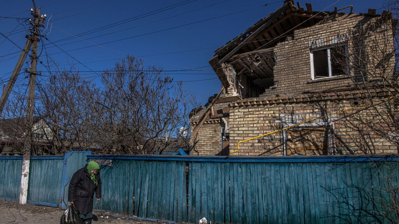Starsza kobieta stoi obok domu zniszczonego przez ostrzał