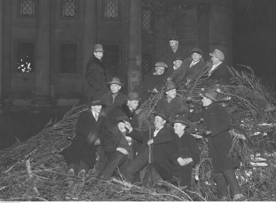 Grupa młodych mężczyzn na stosie niesprzedanych choinek, w oczekiwaniu na rozpoczęcie pasterki. W tle widoczny kościół św. Aleksandra w Warszawie (1926 r.)