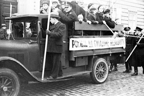Policjanci z dziećmi na samochodzie z puszkami przymocowanych do kijów zbierają datki. Na samochodzie umieszczony transparent z napisem "Policja na Gwiazdkę Bezrobotnym" (NAC, sygnatura 3/1/0/13/1078).