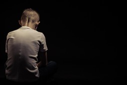 Rear View of a Seated Sad Young Boy Against Black