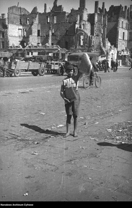 Bosonogi chłopiec sprzedający „Życie Warszawy” w Al. Jerozolimskich. W tle rowerzysta i furmanki, w oddali ruiny kamienicy Lothego przy ul. Marszałkowskiej 100, 1945 r.
