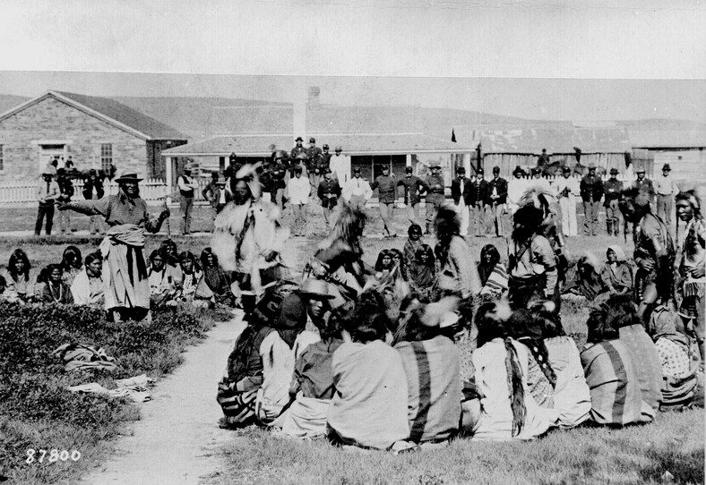 "Szoszoni w Ft. Washakie, rezerwat rdzennych Amerykanów Wyoming. Wódz Washakie (po lewej) wyciąga prawą rękę. Niektórzy tańczą przed przyglądającymi się żołnierzami, 1892 r.