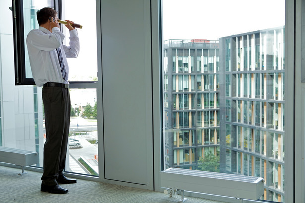 Mężczyzna spoglądający przez lunetę na biurowiec, mat. shutterstock