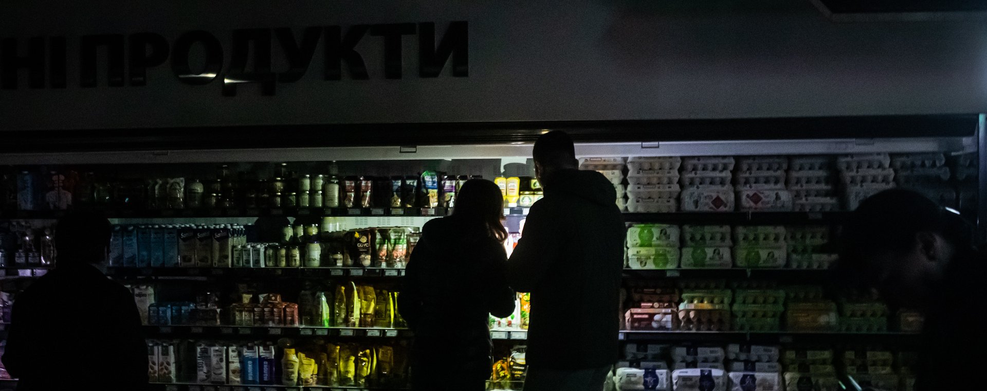 Zakupy w dziale spożywczym supermarketu podczas przerwy w dostawie prądu w Kijowie. Kupujący oświetlają towary telefonami komórkowymi. 27 października 2022 r. 