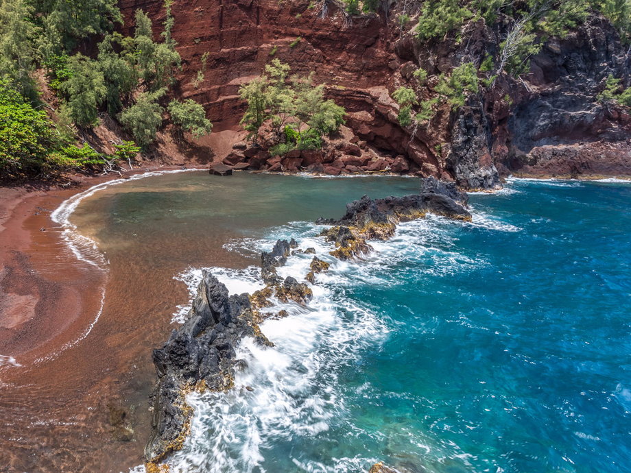 The Red Sand Beach, or Kaihalulu Beach, in Maui, Hawaii, gets its red sands from the volcanic cinder cone surrounding it. The beach, which you can access by descending down a trail, is also protected by a wall of jagged black lava rock, adding to its breathtaking scenery.