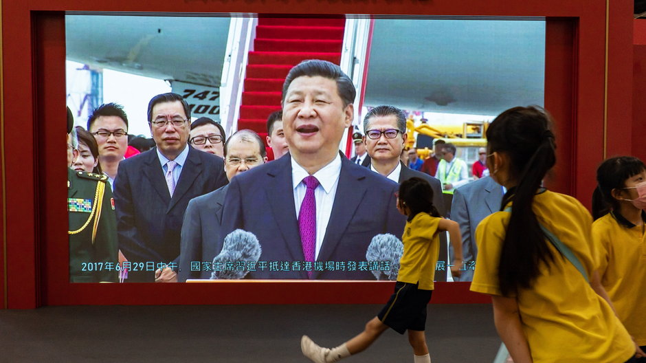 Uczniowie przechodzą przed ekranem telewizora z materiałem filmowym z wizyty Prezydenta Chin, Xi Jinpinga w Hongkongu.