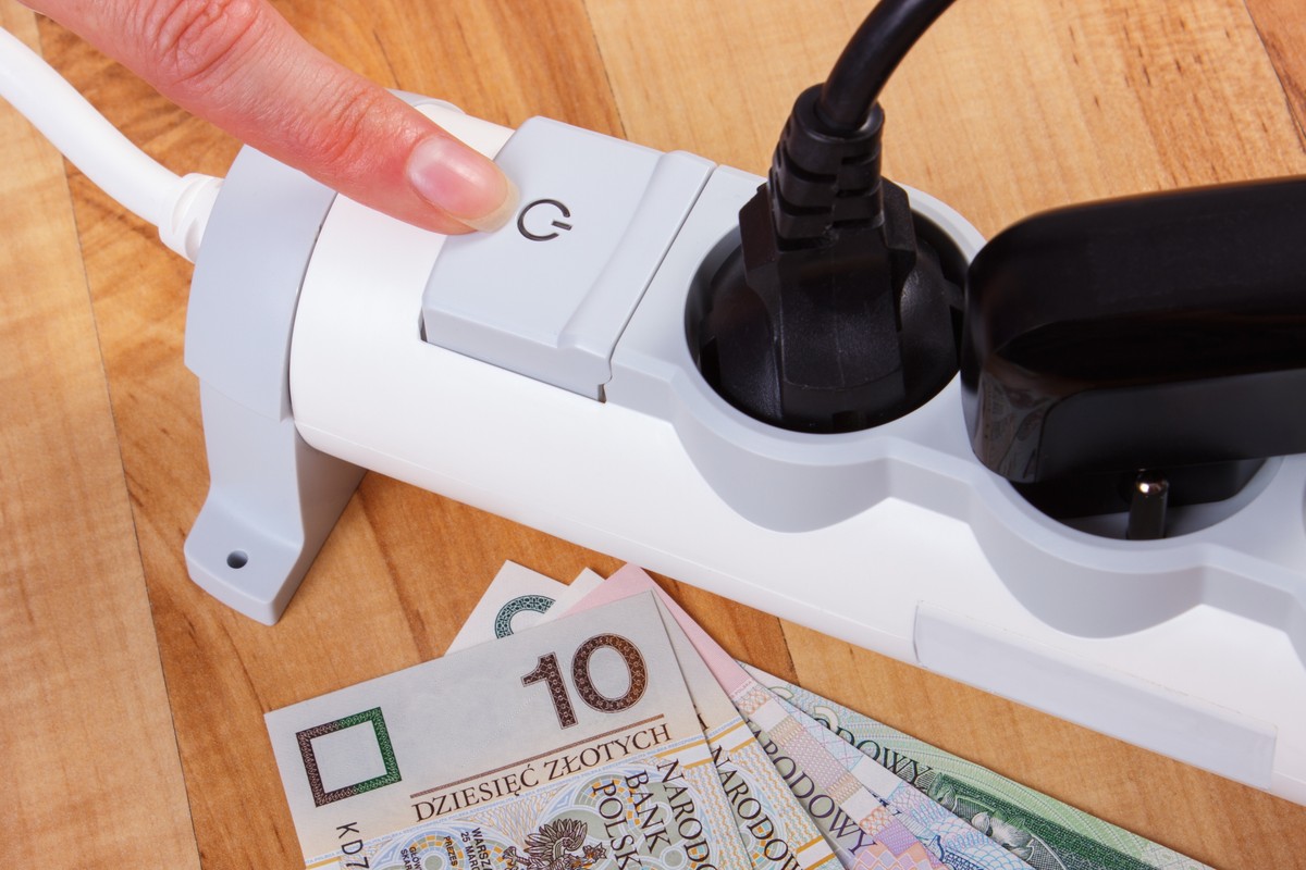Ręka wyłączająca listwę zasilającą z podłączonymi wtyczkami, na stole leżą polskie banknoty – symbol oszczędzania energii i pieniędzy.